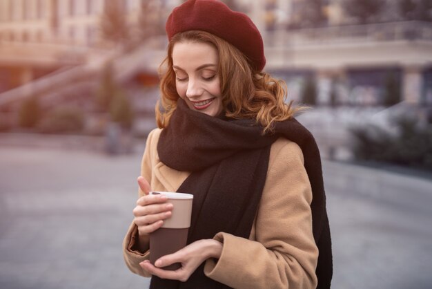 屋外に立っているコーヒーマグを保持している美しいフランス風の若い女性の半身像