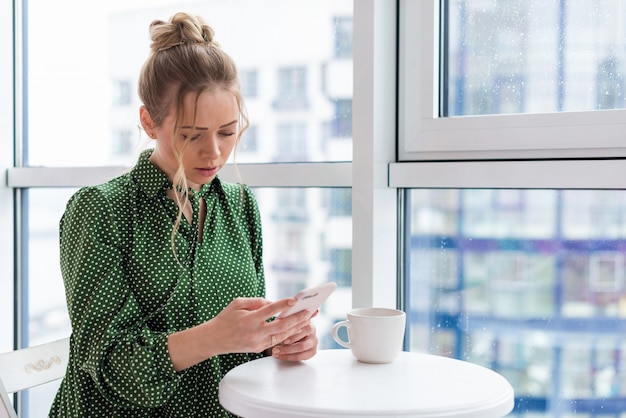 Half-length portrait of blonde girl sits at the table next the window holding a mobile phone and looking at the screen