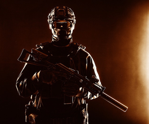 Half-length portrait of special forces soldier, army commando, police tactical team or SWAT fighter with hidden behind mask and glasses face, armed assault rifle with silencer, low key studio shoot