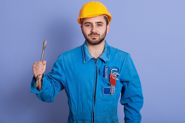 Half length portrait of young bearded manual worker holding wrench tool in one hand and having set of tools in pocket