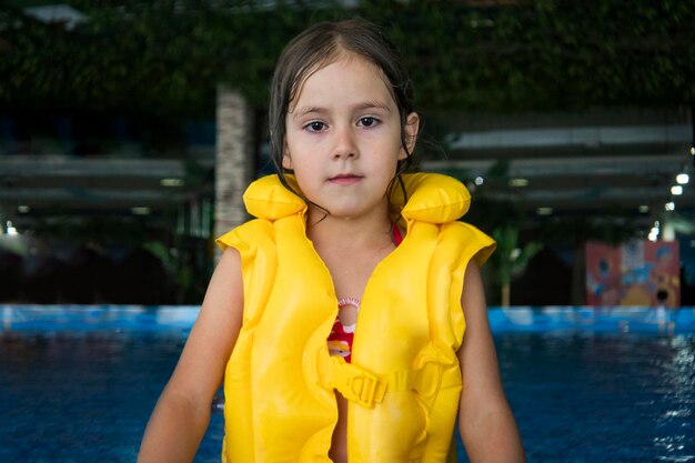 ウォーターパークの背景にある水泳装置でかわいい女の子の半分の長さのショット。小さな女の子は、アクアパークの屋内プールに滞在しているカメラを見ています。アクアパークで泳いだ後の少女