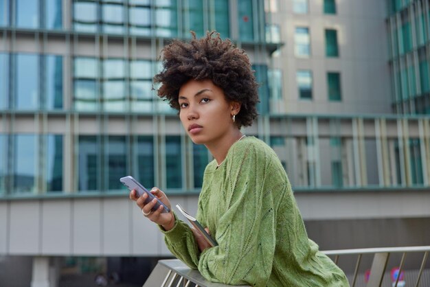 Half length shot of pensive woman with dark curly hair poses at\
urbanity with mobile phone downloads new application for editing\
media files checks notification dressed in casual green jumper