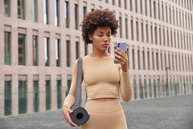 Photo half length shot of sporty woman in beige tracksuit reads text message via smartphone concentrated at screen carries fitness mat prepares for workout