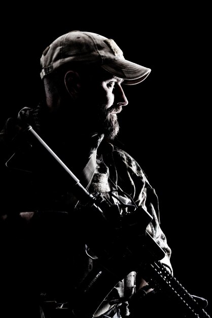 Half length studio contour backlight shot of bearded special forces soldier in uniforms with weapons, portrait on black background.