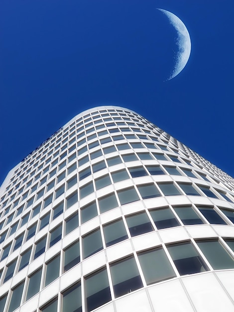 Half a moon over a building skyscraper shining in bright blue sky Below the crescent of quarter moon over a tall modern structural architecture foreboding doomed capitalism or an economical crisis