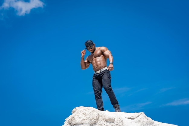 Half naked bodybuilder posing with muscular arms up Background from beautiful white mountains and clear sky Man in sunglasses standing