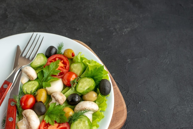 Half shot of fresh black and yellow olives chopped vegetables cutlery set on a white plate on wooden board on black surface