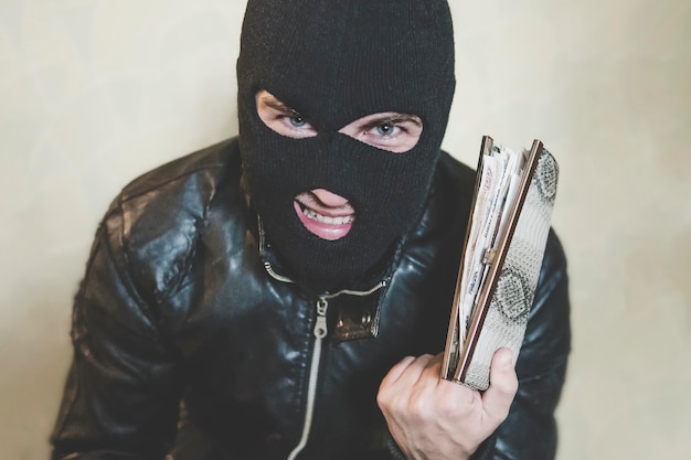 Half shot of gloating pickpocket in black mask looking at the viewer and grinning Selfsatisfied robber with hidden face holds the stolen purse full of money and mocks at the camera Thief plunder