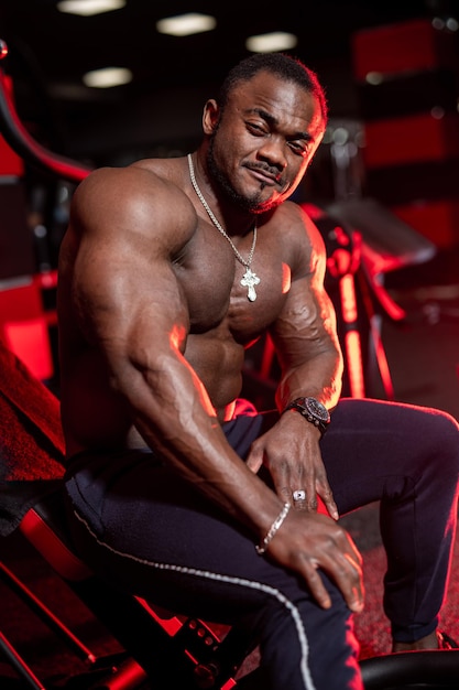 Halfnaakte gerichte Afro-Amerikaanse bodybuilder zittend in moderne donkere sportschool met rood licht.