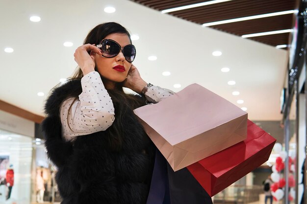 Halfshot van jong aantrekkelijk meisje dat een zonnebril opdoet na het winkelen in het winkelcentrum trendy brunette meisje met papieren zakken wandelen in luxe winkel geld uitgeven in winkels op jacht naar merken