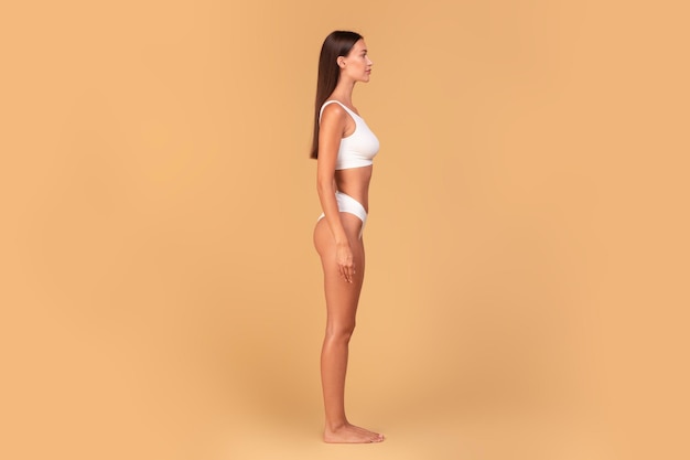 Halfturned slim woman in white underwear posing isolated on beige studio background side view full length copy space
