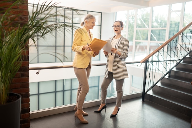 In the hall. Contented Caucasian young businesswoman standing in the hall next to her senior colleague while holding documents in her hands