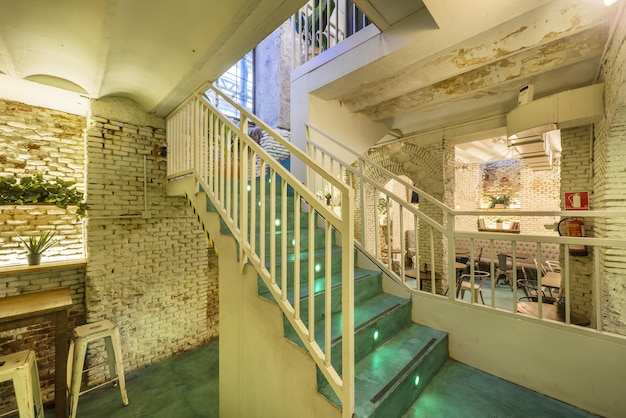 Холл ресторана с деревянной мебелью в индустриальном стиле, окрашенные в белый цвет старые кирпичные стены и лестницы с металлическими перилами и зелеными ступенями с подсветкой