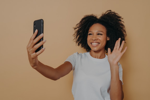 Hallo! mooie vrolijke vrouw met een donkere huid die lacht naar de camera op moderne smartphone, selfie op mobiele telefoon neemt en zich gelukkig voelt, hallo gebarend terwijl ze videogesprek voert, geïsoleerd over studiomuur