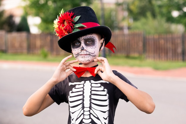 Halloween boy with painted face in costume of skeleton with\
elegant hat and red bowtie on neck standing in front of camera\
against road