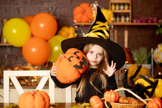 Halloween, a child girl in a witch costume scares in a dark kitchen with pumpkins and a big spider and festive decor during the Halloween celebration