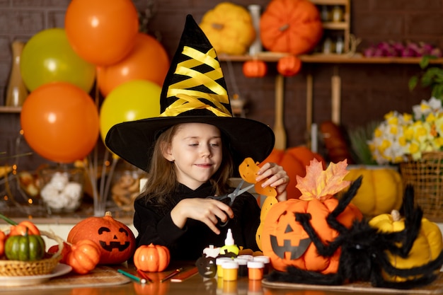 Halloween, a child girl in a witch costume with pumpkins and a big spider in a dark kitchen cuts a festive garland with scissors, smiles, rejoices