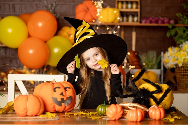 Halloween child a girl in a witch costume with pumpkins and a big spider in a dark kitchen scares or plays with leaves during the halloween celebration