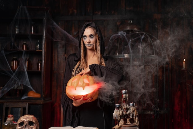 Halloween concept Black witch holding Halloween pumpkin with carved smily face in hand standing dark room