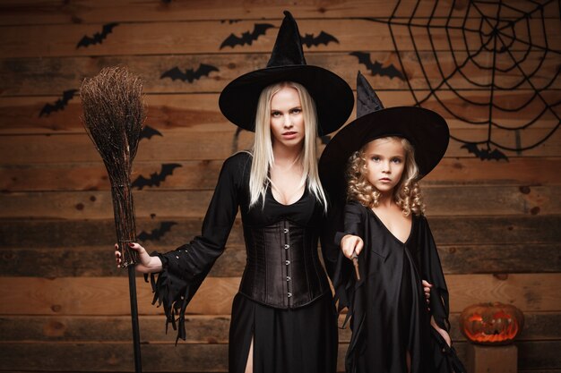 Halloween Concept - cheerful mother and her daughter in witch costumes celebrating Halloween posing with curved pumpkins over bats and spider web on Wooden studio background.