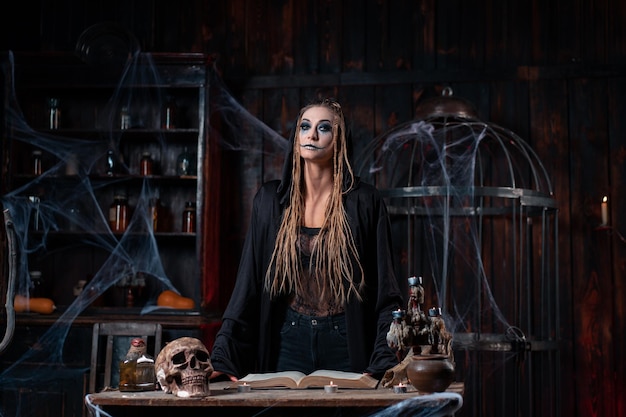 Halloween concept. Witch dressed black hood with dreadlocks standing dark dungeon room use magic book for conjuring magic spell. Female necromancer wizard gothic interior with skull, cage, spider web