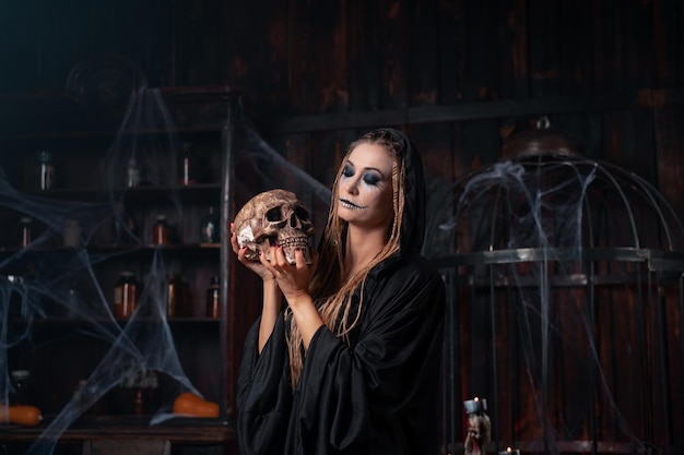 ハロウィーンのコンセプト魔女の肖像画は、背景にケージと暗い部屋に立っているカメラの服を着た黒いフードを探しているドレッドヘアでクローズアップ