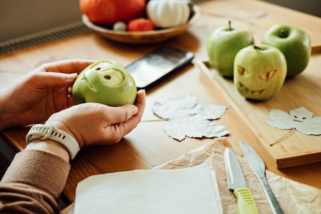 Halloween DIY Fruit ideas Female hands cutting out Halloween green apple with creepy carved face on wooden table at home Healthy Halloween Snacks