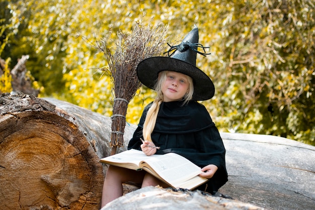 Halloween en heksen. Kind in een heksenkostuum. Het kind zit op een boom met een boek en een toverstaf. Herfst, bos