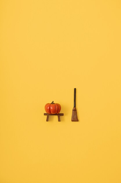 Halloween flat lay concept made of tiny orange pumpkin and witch broom on pastel yellow background. Minimal autumn idea. Creative top view with copy space.