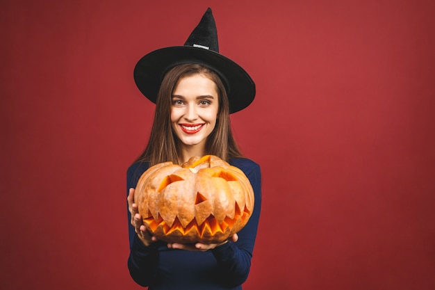 Halloween-Heks met een gesneden Pompoen - die op rode achtergrond wordt geïsoleerd. Emotionele jonge vrouw in Halloween-kostuum.