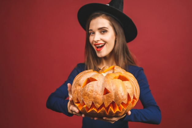 Halloween-Heks met een gesneden Pompoen - die op rode achtergrond wordt geïsoleerd. Emotionele jonge vrouw in Halloween-kostuum.