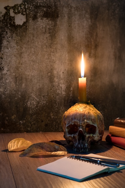 Halloween image with a burning candle on an ancient human skull and books onwood table bac