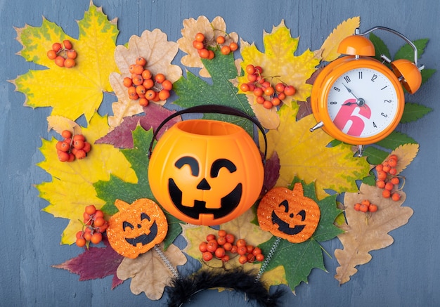 Halloween Jack o Lantern met snoepjes op een kleurrijke herfstachtergrond van gedroogde bladeren, oranje lijsterbessen en een wekker. Halloween-vakantiekaart. Halloween vakantie tijd concept