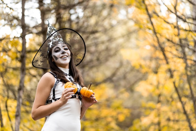 Halloween jong mooi meisje in een witte jurk met make-up op haar gezicht in een heksenhoed die zich voordeed in het herfstbos Hij houdt twee kleine pompoenen in zijn handen en glimlacht De dag van de doden