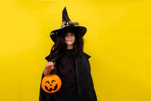 Halloween kid holds candy bowl in his hand jack o lantern and waiting for sweets Child in black sorcerer39s costume Witch yellow background Boy wizard Copy space Trick or treating