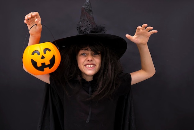 Halloween kid raises his hands up holding jack o lantern and makes faces to scare and get sweets Child in black sorcerer39s costume Witch black background Boy wizard Copy space Trick or treating