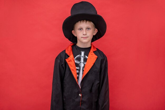 Halloween kids A boy in a skeleton costume holds a jack o lantern Red background Copy space A child in a black coat hat holds a halloween pumpkin Place for text