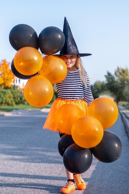 Foto bambini di halloween ritratto sorridente ragazza con cappello da strega con palloncini arancioni e neri bambini divertenti in costumi di carnevale all'aperto