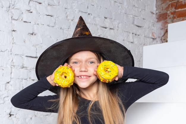 Halloween-kinderen een lachend blond meisje in een heksenkostuum met gele donuts in haar handen zittend op de trap