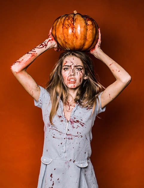 Halloween-meisje met een gesneden pompoen. Portret van een vrouw met make-up bloed op haar gezicht. Sexy vrouw draagt blauwe drees met bloed. Ze heeft pompoen in handen boven het hoofd.