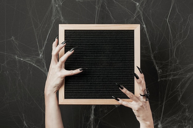 Halloween mock up Letter board in hands with long black nails and spider ring on black background with spiderweb Front view