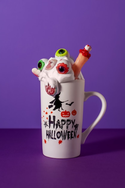 Halloween monster shake in lange mok op paarse achtergrond. Slagroom met ogen, vinger, hersenen en schedelsnoepjes. Griezelig drankje.