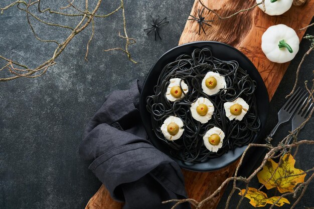 Halloween party Italian black pasta decorated horror olives like eyes on black plate on old dark table background Monster face from pasta Halloween decorations and Halloween food concept Mock up