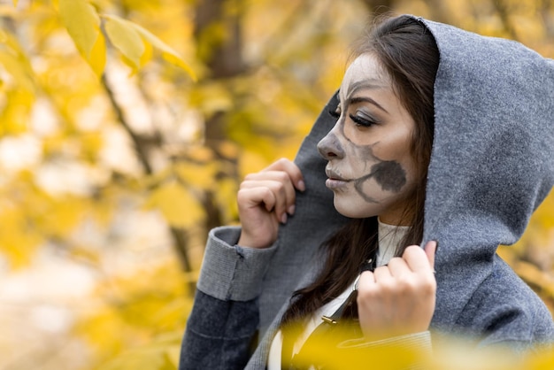 Halloween portrait A young beautiful girl in a white dress with makeup on her face in a hooded mantle creepily stands in the autumn forest The day of the Dead