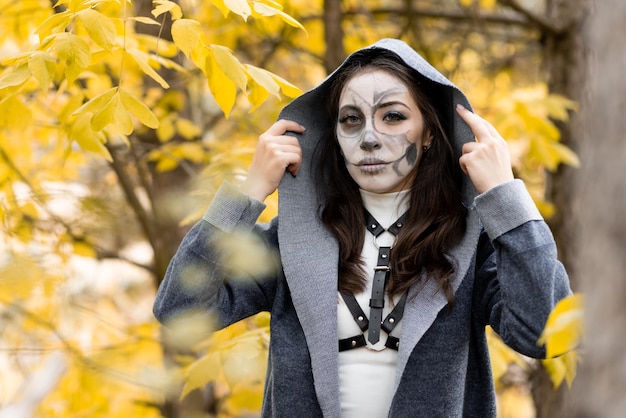 Halloween-portret Een jong mooi meisje in een witte jurk met make-up op haar gezicht in een mantel met capuchon staat griezelig in het herfstbos De dag van de doden