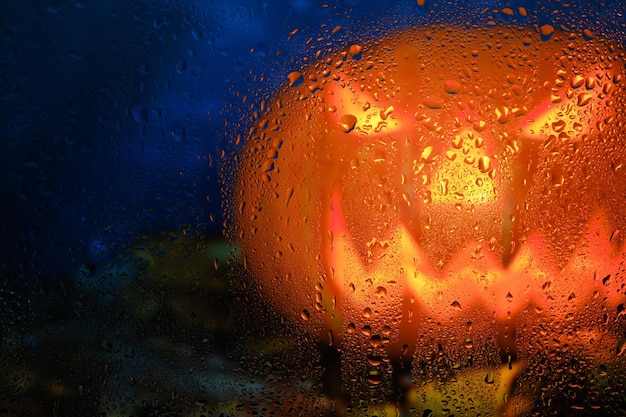 雨滴で濡れた秋の窓の後ろに燃えるような目を持つハロウィーンのカボチャ。