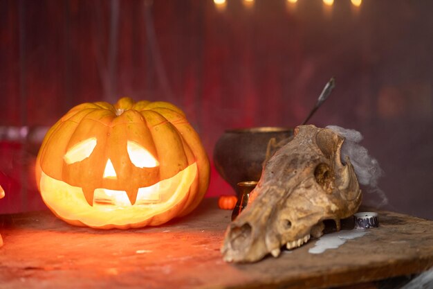 Хэллоуин. Страшная тыква Хэллоуина с вырезанным лицом на столе в темной комнате с человеческим черепом и черепом животного. Скопируйте место для текста. Жуткая атмосфера.