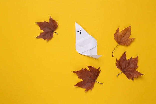Halloween-thema Origami-spook met gevallen herfstbladeren op een gele achtergrond Bovenaanzicht
