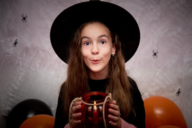Foto halloween voor kinderen een lachend meisje in een heksenhoed met een glazen pot in de vorm van een pompoen.