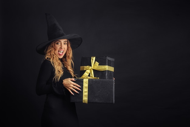 Halloween witch with a black hat on a black wall holding gift boxes